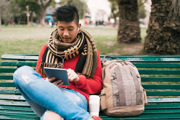 Portrait of young Asian man using his digital tablet with earphones while sitting in a bench outdoors. Technology concept. - Fotografie, Obrázek