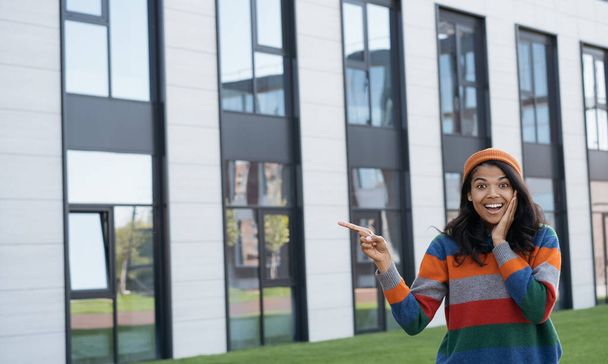 Feliz mujer afroamericana emocionada usando ropa casual y sombrero naranja señalando con la mano en el espacio de copia, de pie al aire libre - Foto, imagen