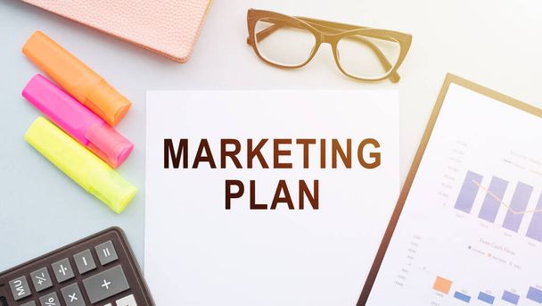 Paper with text marketing plan on table with colorful markers, glasses, financial graphs, calculator and notebook - Фото, зображення