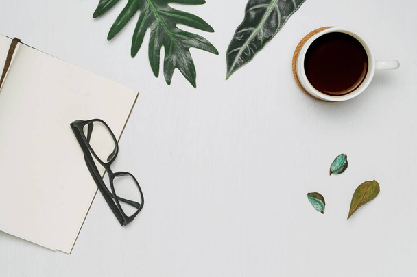 Xícara de café e caderno com óculos em fundo de madeira branca com folhas de monstera tropicais. Deitado plano mínimo, vista superior. - Foto, Imagem