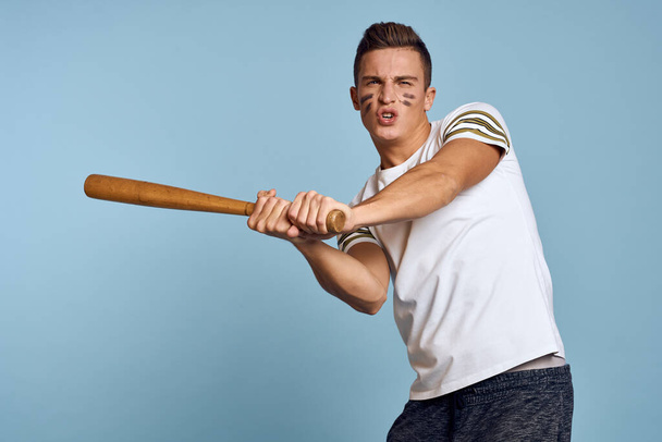 τύπος του μπέιζμπολ με ένα ρόπαλο στο χέρι του σε ένα μπλε φόντο και ένα χρώμα μάχης στο πρόσωπό του μακιγιάζ μοντέλο t-shirt - Φωτογραφία, εικόνα