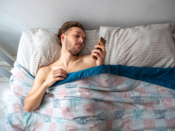 Νεαρός καυκάσιος άνδρας κοιτάζει το τηλέφωνό του στο κρεβάτι το πρωί - Φωτογραφία, εικόνα
