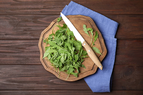 Cutting board with fresh arugula leaves and knife on wooden table, flat lay - Фото, зображення