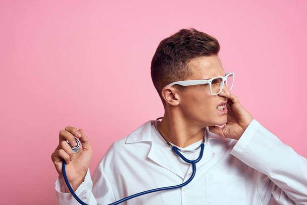 Pembe bir arka planda steteskop ve yüzünde gözlük olan tıbbi önlüklü bir erkek doktor. - Fotoğraf, Görsel