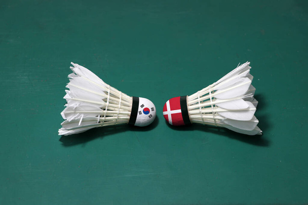 Dwa używane wahadłowce na zielonym piętrze sądu w Badminton z obiema głowami. Jedna głowa namalowana flagą Korei Południowej i jedna głowa namalowana flagą Danii, koncepcja konkursu badmintona. - Zdjęcie, obraz
