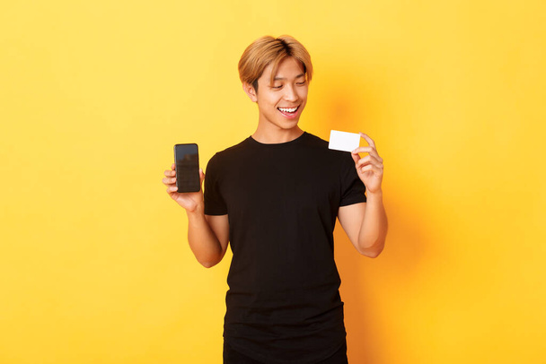 bello soddisfatto asiatico ragazzo in possesso di carta di credito e mostrando smartphone schermo sorridente soddisfatto in piedi giallo sfondo - Foto, immagini