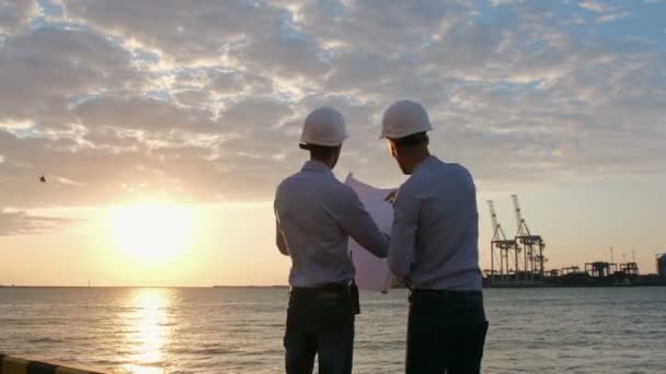 Engineers in white safety helmets discuss a construction plan with cranes in the background at dawn. The man points with his hand to the drawing and then into the distance. - Footage, Video