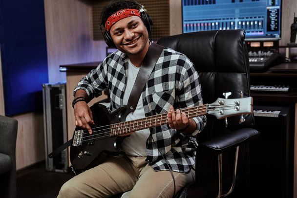 Portrait of happy young man, male artist in headphones smiling aside, playing guitar while sitting in recording studio - Photo, Image