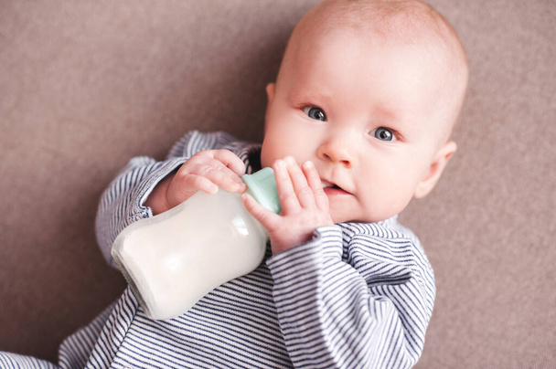 Newborn child drinking milk holding bottle lying in bed. Looking at camera. Healthy nutrition.  - Photo, Image