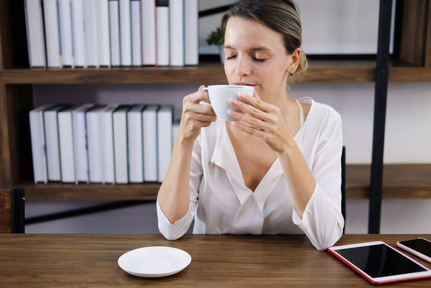 Hymyilevä valkoihoinen nainen tyytyväinen kupin kuumaa kahvia aamulla auringonnousun kahvilassa. Hän rentoutuu tabletin kanssa kahvilassa toimistossa. Liikenaisen elämäntapa ja teknologian käsite - Valokuva, kuva