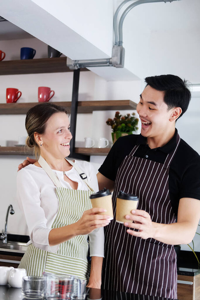 Sorrindo jovem barista casal ama o homem asiático e mulher caucasiana está abraçando e segurando copos de café quente juntos. Iniciar Café e café conceito de negócio. - Foto, Imagem