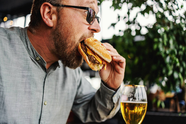 Homem com fome barbudo de meia idade sentado no restaurante e comendo delicioso hambúrguer. - Foto, Imagem