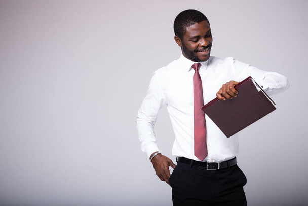 Ένας Αφροαμερικάνος μεσίτης στέκεται με ένα φάκελο στα χέρια του, κοιτάζει το ρολόι του και περιμένει τον πελάτη του. - Φωτογραφία, εικόνα