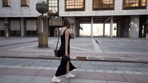 Eine junge Frau im langen schwarzen Kleid macht ein Foto von der Stadt und lächelt. Handeln. Schöne junge Touristin, die mit einer professionellen Kamera im Hintergrund der Gebäude des Geschäftsviertels spaziert - Foto, Bild
