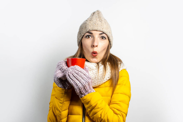 Mladá žena ve žlutém saku a klobouku drží sklenici kávy nebo čaje na světlém pozadí. Emoční smích, překvapení, polibek. Koncept zima, podzim, kavárna. - Fotografie, Obrázek