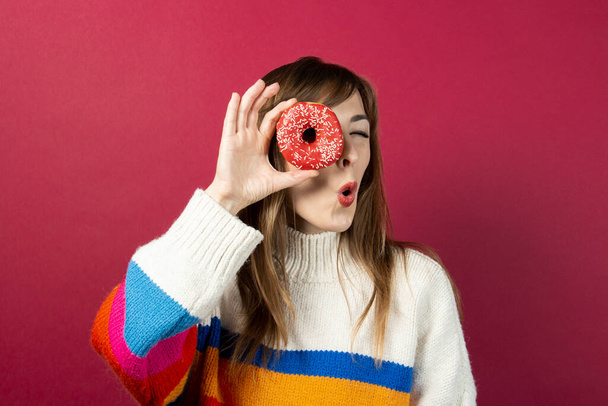 νεαρή κοπέλα σε ένα πλεκτό πουλόβερ, κρατά ένα ντόνατ στο χέρι της και κοιτάζει με έκπληξη το άνοιγμα του ντόνατ σε ένα μπορντό φόντο. Μπάνερ. - Φωτογραφία, εικόνα