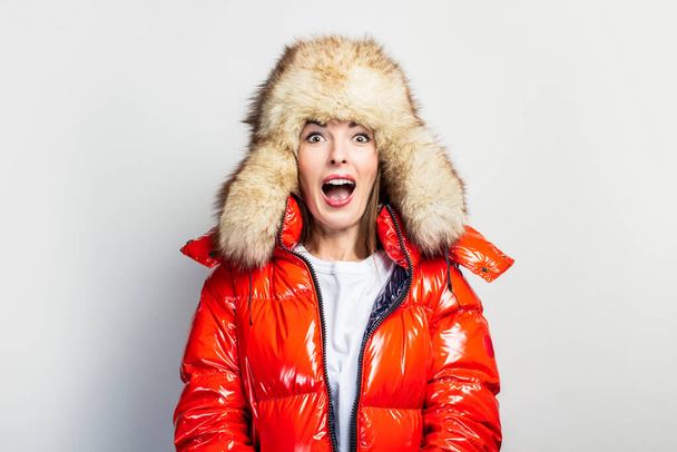 young girl in a red jacket and a fur hat, screaming with open eyes on a light background. Concept shock, surprise. - Fotoğraf, Görsel