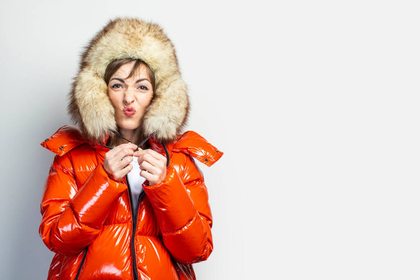 young girl in a red jacket and a fur hat, makes a kiss with her lips, puckered on a light background. Concept emotions, gestures, feelings. - Photo, Image