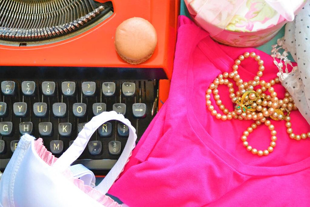 Vista aérea de una máquina de escribir con un macaron en la parte superior rodeado de cosas como un sujetador, una camiseta, collares de perlas, una bufanda estrella o una caja de regalo - Foto, imagen
