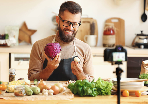 Счастливый бородатый мужчина в очках улыбается и демонстрирует спелые овощи во время съемки видео для приготовления видеоблога на кухне у дома - Фото, изображение
