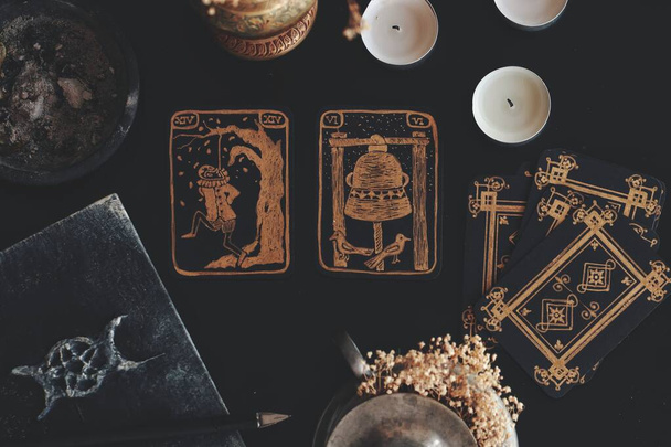 Flat lay of black and gold tarot card art. Hand made, not real diy vintage antique looking tarot cards on black table. Messy wiccan witch altar with grimoire, flowers, candles on it. The hanged man. - Photo, Image
