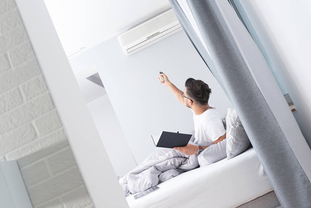 Man switching on air conditioner relaxing on bed - 写真・画像