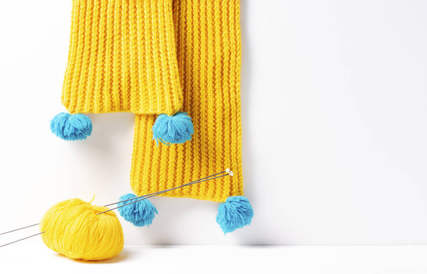 A yellow knitted scarf hangs, next to it lies a yellow yarn with knitting needles on a white background. Close-up, selective focus on the needles. Horizontal orientation, copy space. - Foto, afbeelding