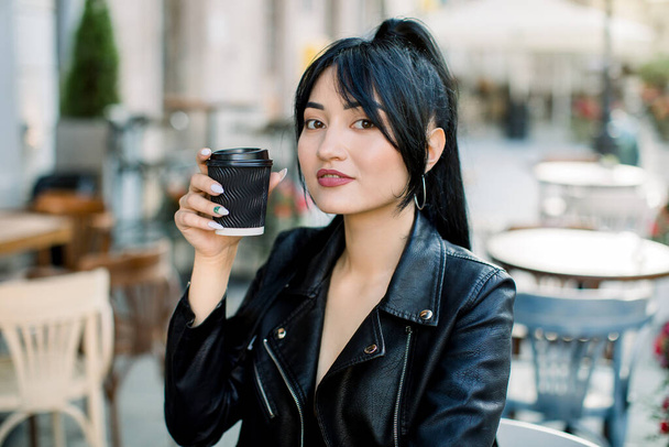 Close-up portrait of attractive stylish smiling Asian girl with brunette ponytail hair, wearing casual black leather jacket, holding paper-cup coffee, sitting outdoors in city cafe - Фото, зображення