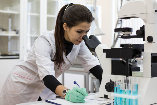 A female chemist looks through a microscope and makes notes in a journal. Scientific research using a microscope in the laboratory. Woman scientist. - 写真・画像