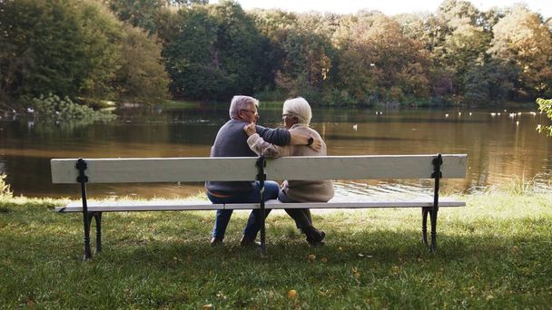 Ηλικιωμένοι γκριζομάλλης ζευγάρι κάθεται στον πάγκο κοντά στο ποτάμι την ημέρα του φθινοπώρου. Ειδύλλιο και γήρας - Φωτογραφία, εικόνα