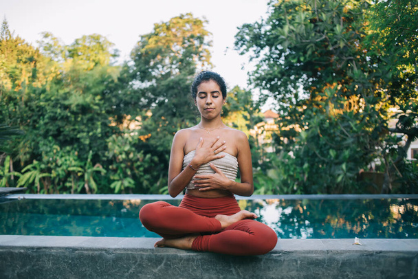 Peaceful young ethnic woman in activewear sitting at poolside in Lotus pose and meditating with closed eyes during yoga session in garden - Foto, immagini