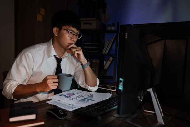 Ελκυστικός νεαρός Ασιάτης επιχειρηματίας συγκεντρώθηκε δουλεύοντας μέχρι αργά υπερωρίες στη δημοσίευση στο γραφείο του στο σκοτεινό μοντέρνο γραφείο τη νύχτα, Υπερφορτωμένος έννοια  - Φωτογραφία, εικόνα