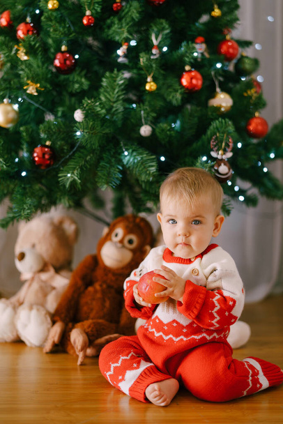 Il bambino con una tutina di Natale rossa e bianca tiene una mela davanti a un albero di Natale. - Foto, immagini