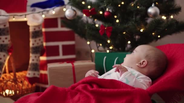 Vánoce, zima, Nový rok, Celebration, rodina, Dětství koncept - šťastný legrační dítě novorozenec ve vánočním oblečení úsměv u vánočního stromečku v prázdninách. Dítě v houpacím křesle zakryté měkkou teplou přikrývkou - Záběry, video