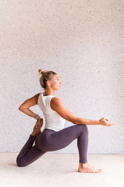 Mode de vie sain. Jeune femme séduisante pratiquant le yoga, portant des vêtements de sport, chemise blanche et pantalon violet, intérieur pleine longueur, fond gris - Photo, image