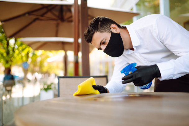Dışarıdaki kafede masaları dezenfekte ederken koruyucu maske ve eldiven takan garson. COVID-19 'u önlemek için masanın temizlenmesi ve dezenfekte edilmesi. - Fotoğraf, Görsel