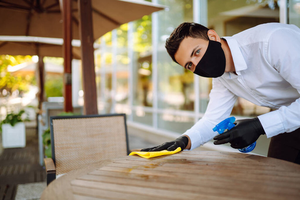 Waiter wearing protective face mask and gloves while disinfecting tables at outdoor cafe. Cleaning and disinfection of table to prevent COVID-19. - Foto, Bild