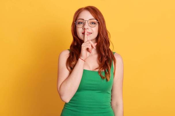 Closeup photo of attractive pretty cute nice glad charming lady holding forefinger near mouth isolated over yellow background, red haired female in green t shirt and spectacles. - Фото, изображение