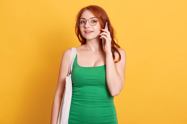 Happy smiling beautiful woman with red hair posing against yellow wall and talking via phone, holding cotton bag, wearing green attire, looks at camera, - Foto, imagen