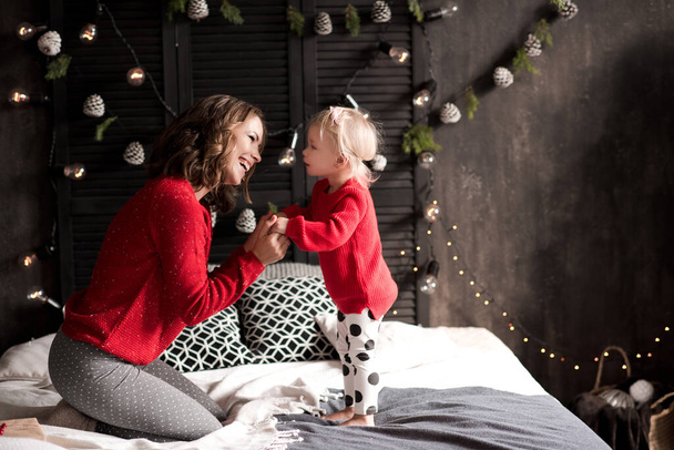 Mother holding baby girl 1-2 year old wearing red knitted sweaters over Christmas decor in room. Winter holiday season. Motherhood. Xmas. - Photo, Image