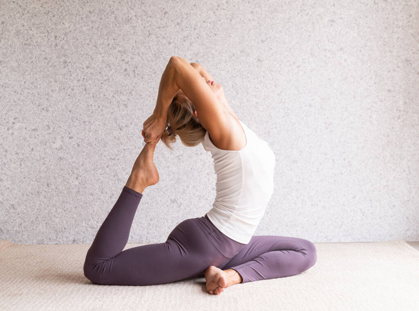 Estilo de vida saludable. Mujer atractiva joven practicando yoga, usando ropa deportiva, camisa blanca y pantalones morados, interior de cuerpo entero, fondo gris - Foto, imagen