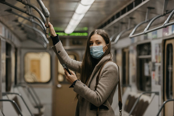 コロナウイルスの拡散を避けるために医療用フェイスマスクの女性が地下鉄の車の中でスマートフォンを持っています。COVID-19に対する外科用マスクの女の子は電車の中で彼女の携帯電話でニュースをスクロールしています. - 写真・画像
