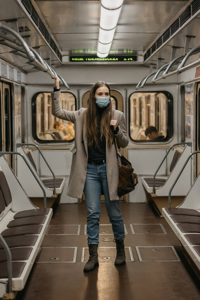 Una mujer con una máscara facial para evitar la propagación del coronavirus se aferra a la barandilla en un vagón del metro. Una chica con el pelo largo en una máscara quirúrgica contra COVID-19 está de pie en un tren de metro. - Foto, Imagen