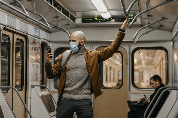 Человек с бородой в медицинской маске для лица, чтобы избежать распространения коронавируса, использует смартфон в вагоне метро. Лысый парень в хирургической маске против COVID-19 держит мобильник в метро - Фото, изображение