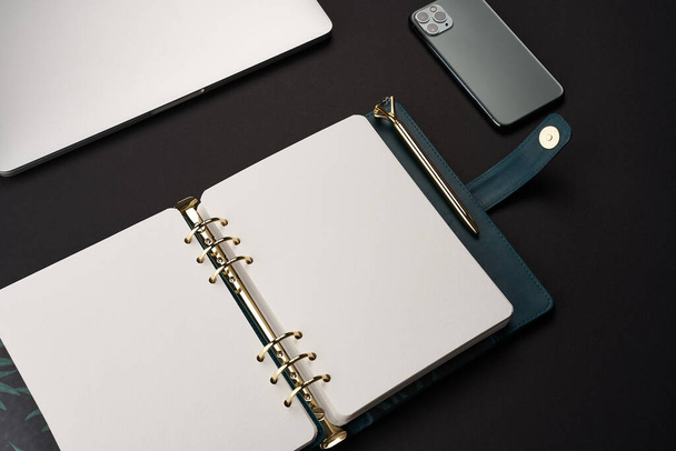 Black desk with open green handmade notebook with gold pen and gray laptop and smartphone - Foto, immagini