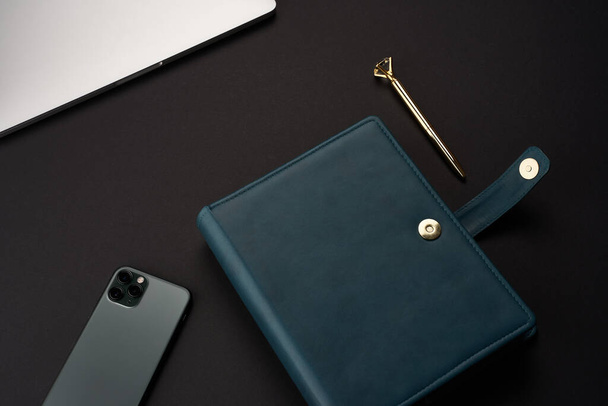 Black desk with green handmade notebooks, gray laptop and smartphone and gold pen - Foto, immagini