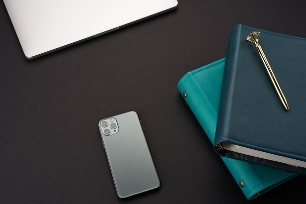 Black desk with green handmade notebooks, gray laptop and smartphone - Foto, immagini