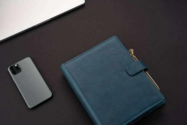 Black desk with green handmade notebooks, gray laptop and smartphone and gold pen - 写真・画像