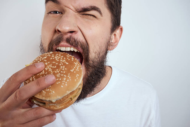 Egy férfi hamburgert eszik könnyű háttérrel fehér pólóban kivágott szemmel közelről éhség gyorsétterem - Fotó, kép