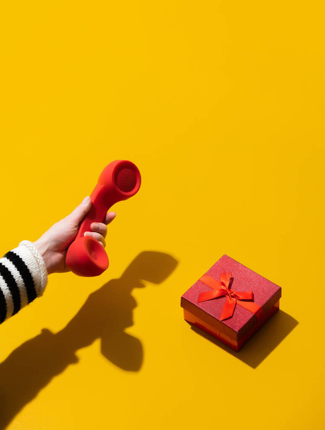 Female hand holds red handset near gift box on yellow background  - Zdjęcie, obraz